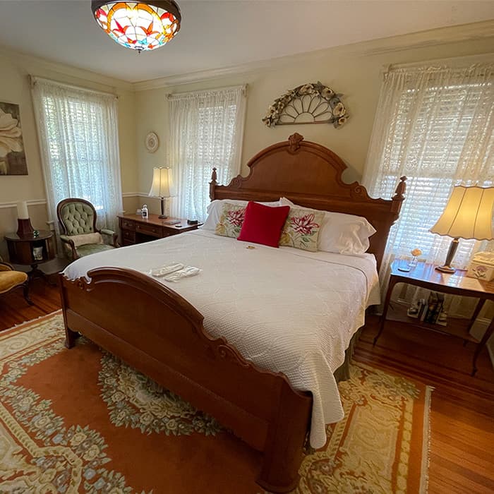 A cozy bedroom featuring a wooden bed, decorative pillows, and vintage lamps, with light streaming through sheer curtains.