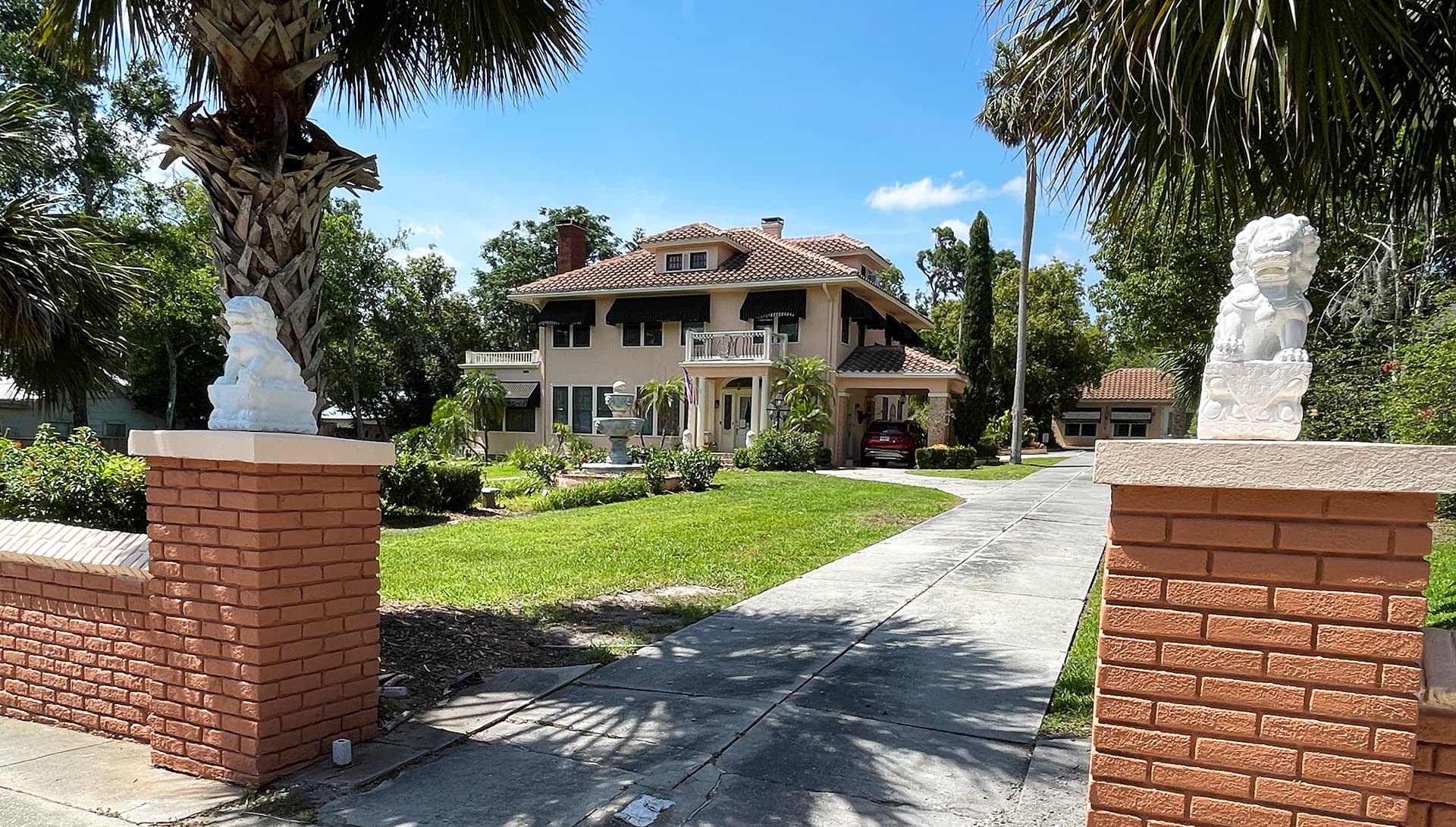 A large, two-story house with a tile roof, flanked by palm trees and lion statues at the entrance.